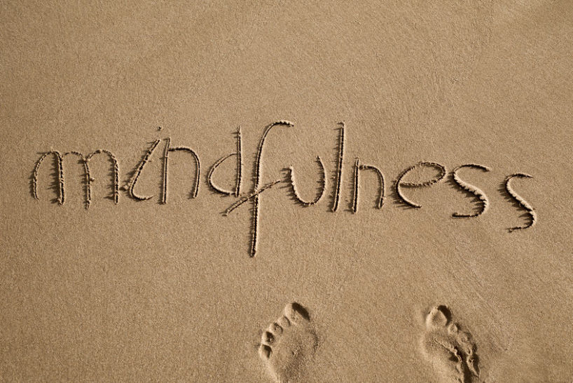 high-angle shot of the word mindfulness written in the sand of a beach and a pair of human footprints